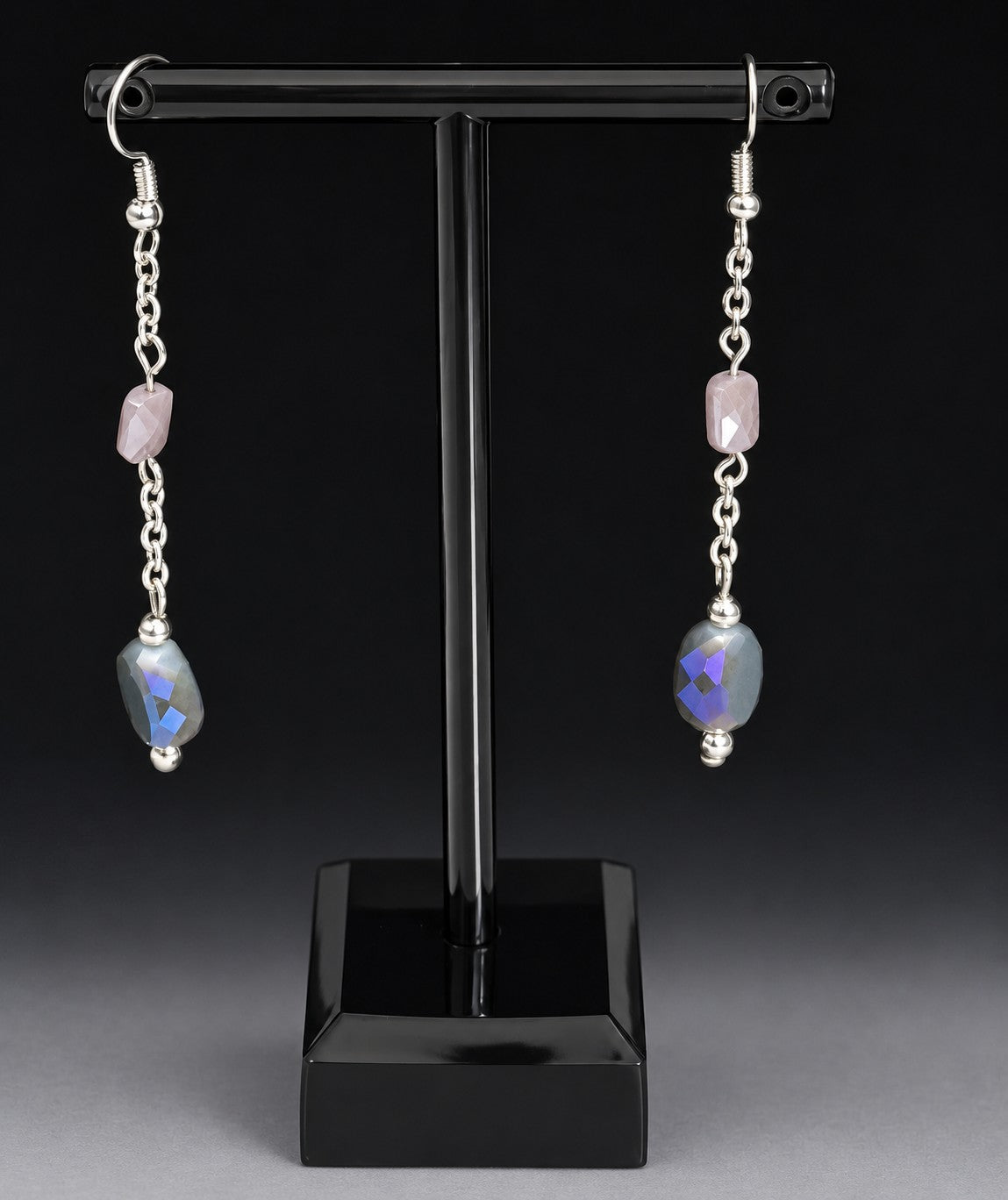 A pair of silver dangle earrings displayed on a black T-stand against a dark studio background. Each earring has a small pink stone, a silver chain, and a larger faceted blue-purple stone with a triangular cutout.