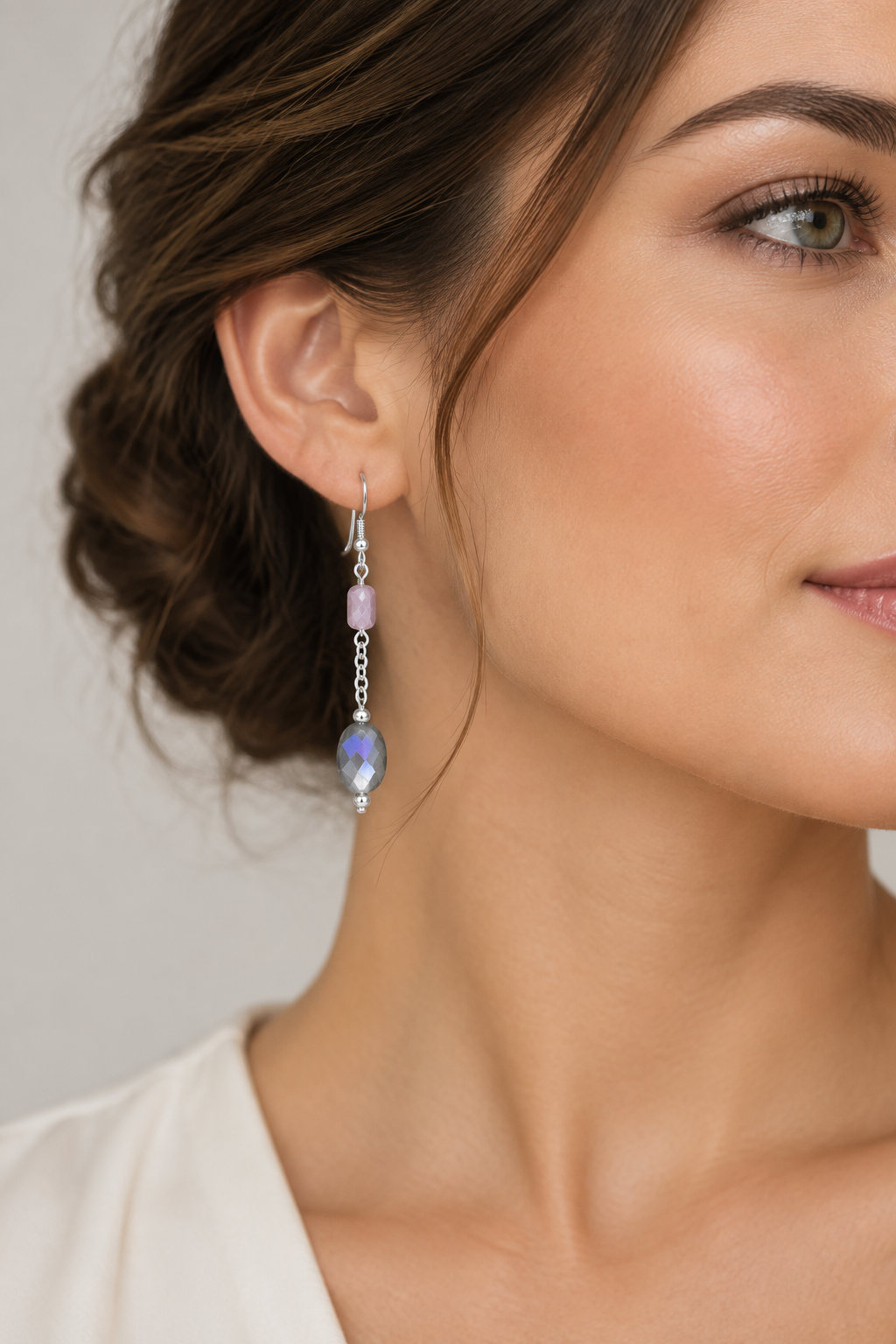 A close-up lifestyle shot of a woman wearing a silver-toned dangle earring with a faceted lavender-purple bead and a small pink accent bead, shown against a soft light gray background.
