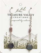A pair of dangle earrings displayed on a branded jewelry card. Each earring has silver-tone hooks and chains, a small dusty pink faceted bead, a dark gray round bead, and a larger dark blue-gray iridescent oval bead.