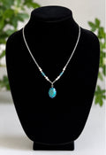 A silver chain necklace with a teardrop-shaped blue-green pendant and small turquoise beads, displayed on a black velvet jewelry bust.