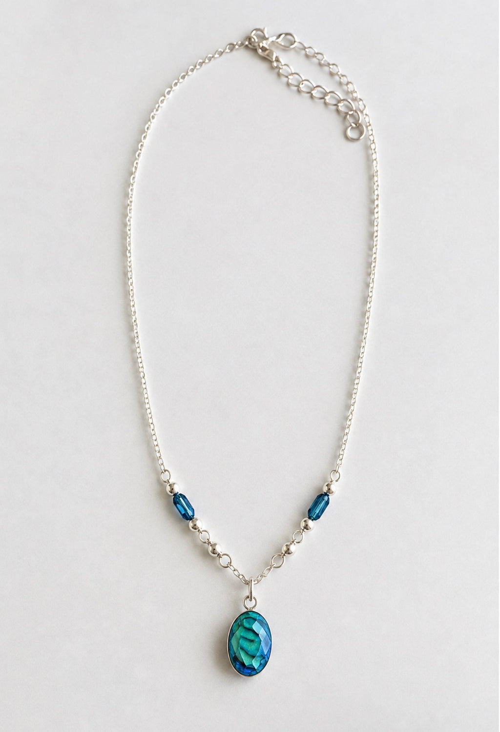 A silver chain necklace featuring a teardrop-shaped blue pendant and two smaller blue beads, displayed on a light gray background.