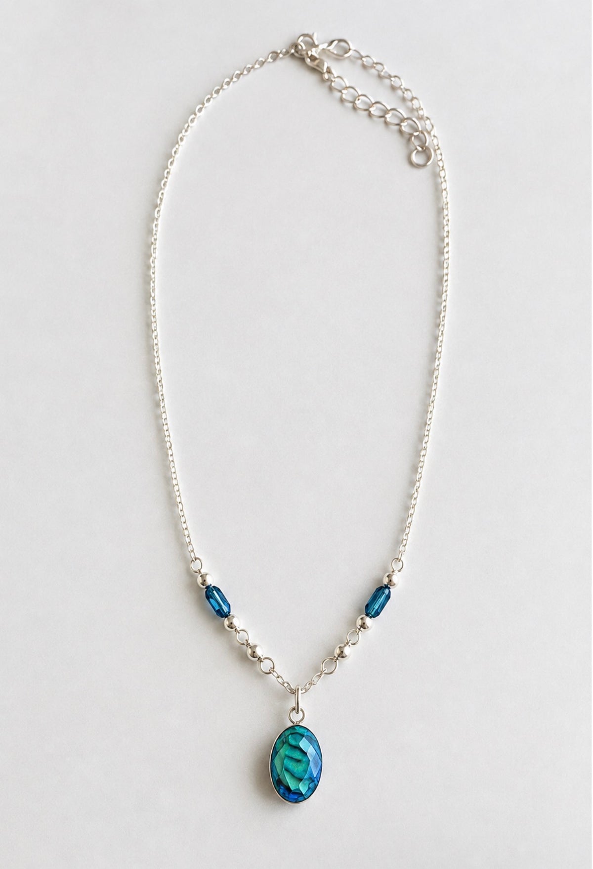 A silver chain necklace featuring a teardrop-shaped blue pendant and two smaller blue beads, displayed on a light gray background.